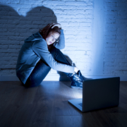 Image of a teen girl sitting in a dark room on the floor in front of the laptop looking worried