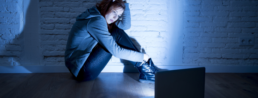 Image of a teen girl sitting in a dark room on the floor in front of the laptop looking worried