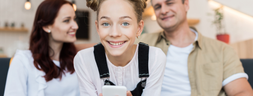 Image of a smiling girl with a smartphone in her hands and her parents in the background blurry and smiling