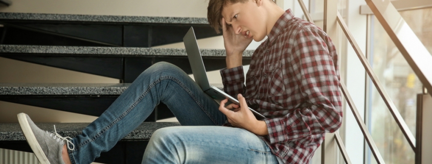 Image of a teen boy sitting on the stairs a looking worrying at his laptop