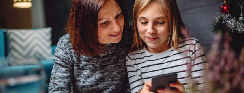Image of a girl smiling while looking at her smartphone and showing it to her mother