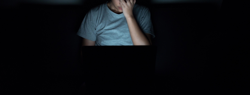 Image of a young boy sitting in the dark in front of a laptop and holding his head in worry