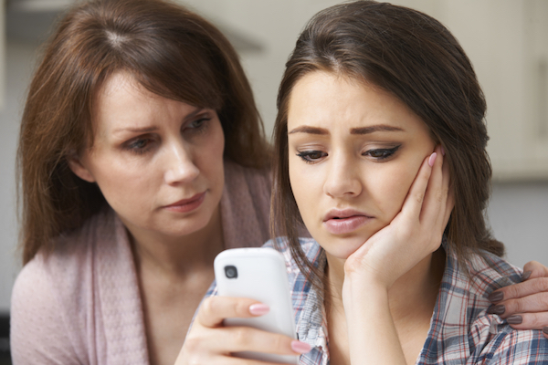 Image of a teen girl looking worried while looking at her smartphone and her mother sitting next to her and hugging her in comfort