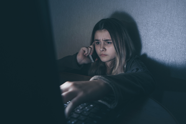 Image of a worried girl in a dark room talking on her phone and typing on the laptop