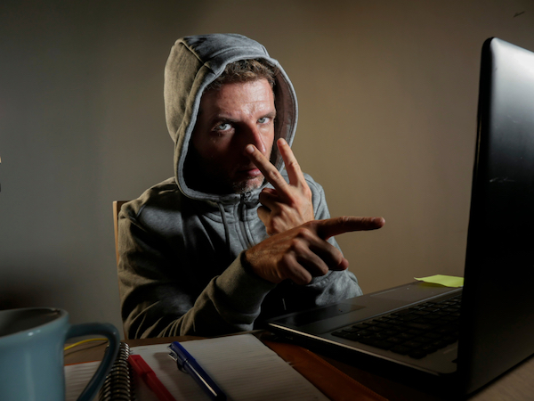 Image of an a man in a hoodie pointing at his eyes with his left index and middle finger and pointing at the laptop with his right index finger