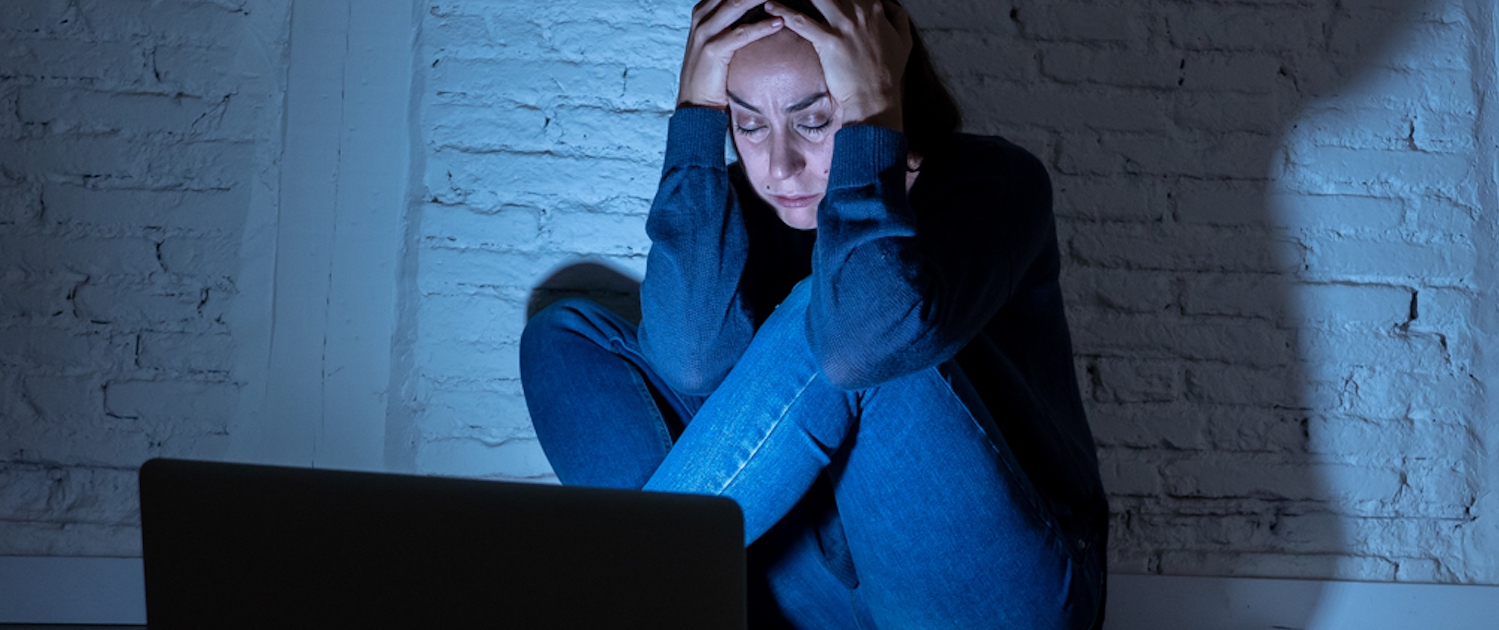 Image of a teen girl sitting in a dark room on the floor in front of the laptop holding her head in worry