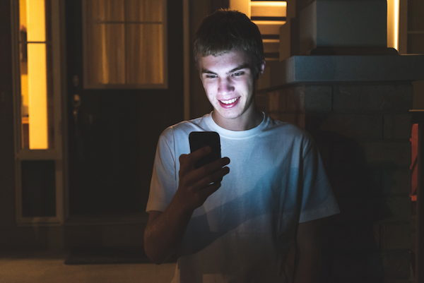 Image of a young teen boy outside of a house looking at his mobile phone in the evening and smiling